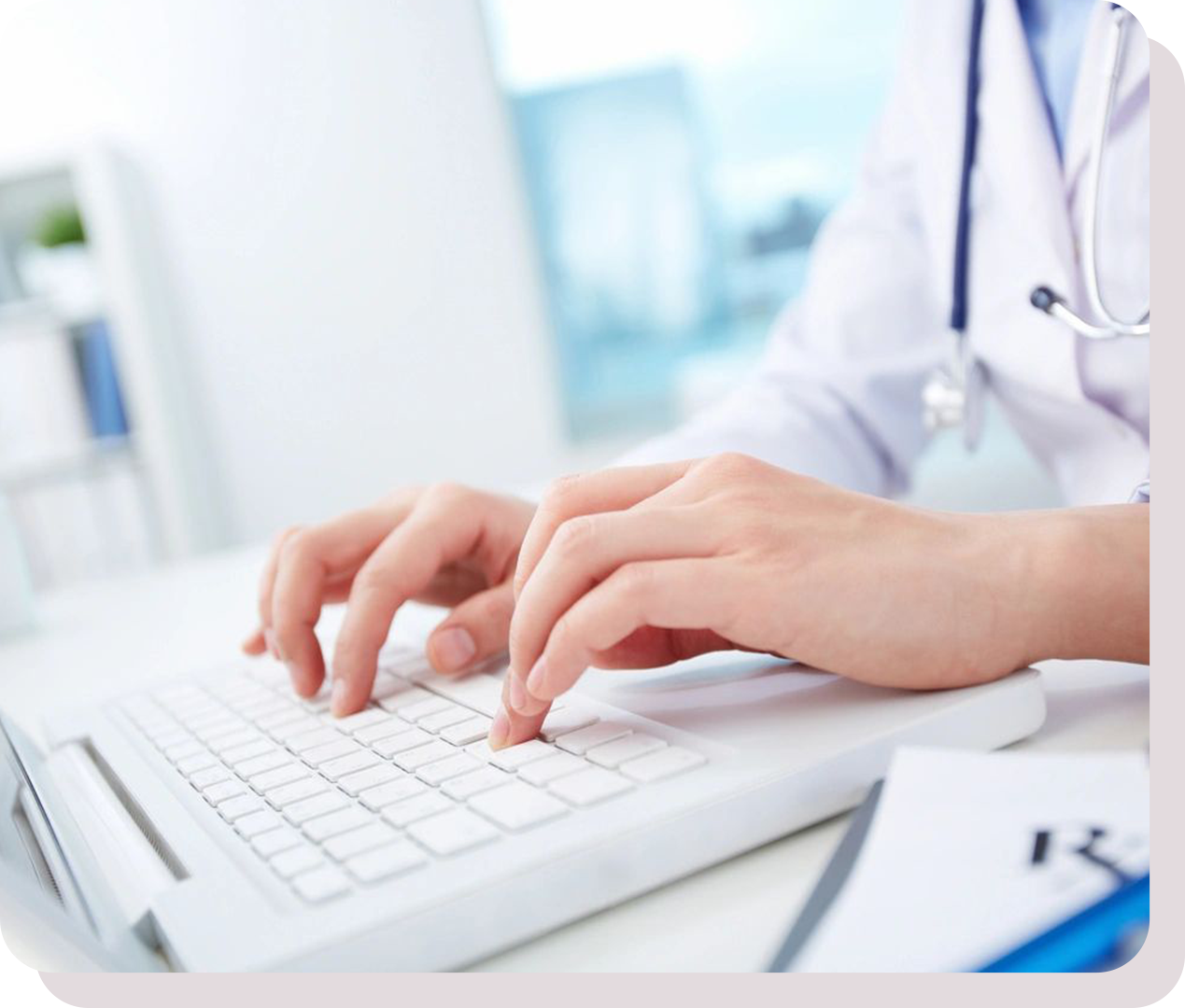 A person in a white coat types on a laptop keyboard. A stethoscope is draped around their neck, and papers are visible on the desk.