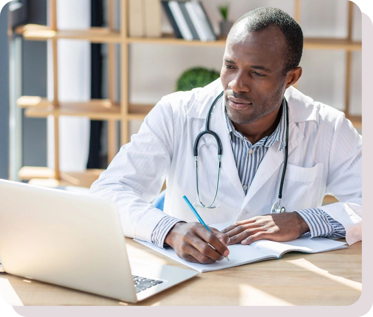 A doctor wearing a white coat and stethoscope writes in a notebook while looking at a laptop on a desk.