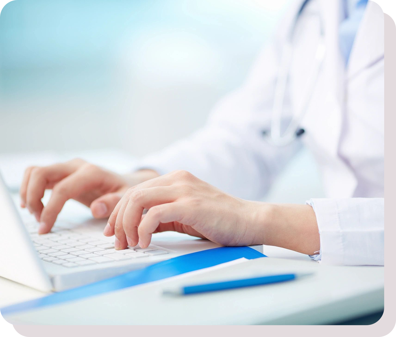 A person in a white medical coat is typing on a keyboard, with a blue pen and documents on the desk.