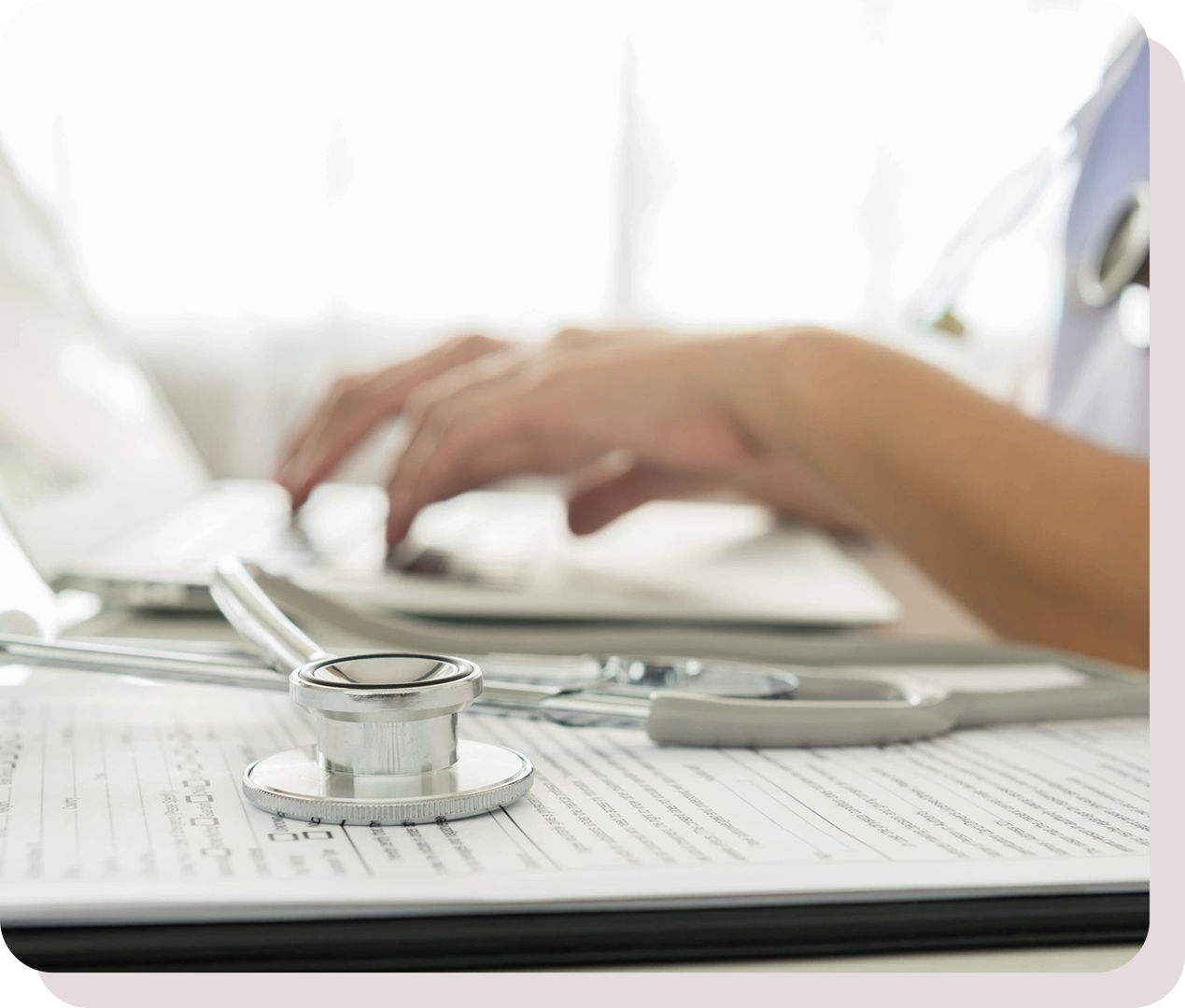 A stethoscope on a medical form with a person typing on a laptop in the background.