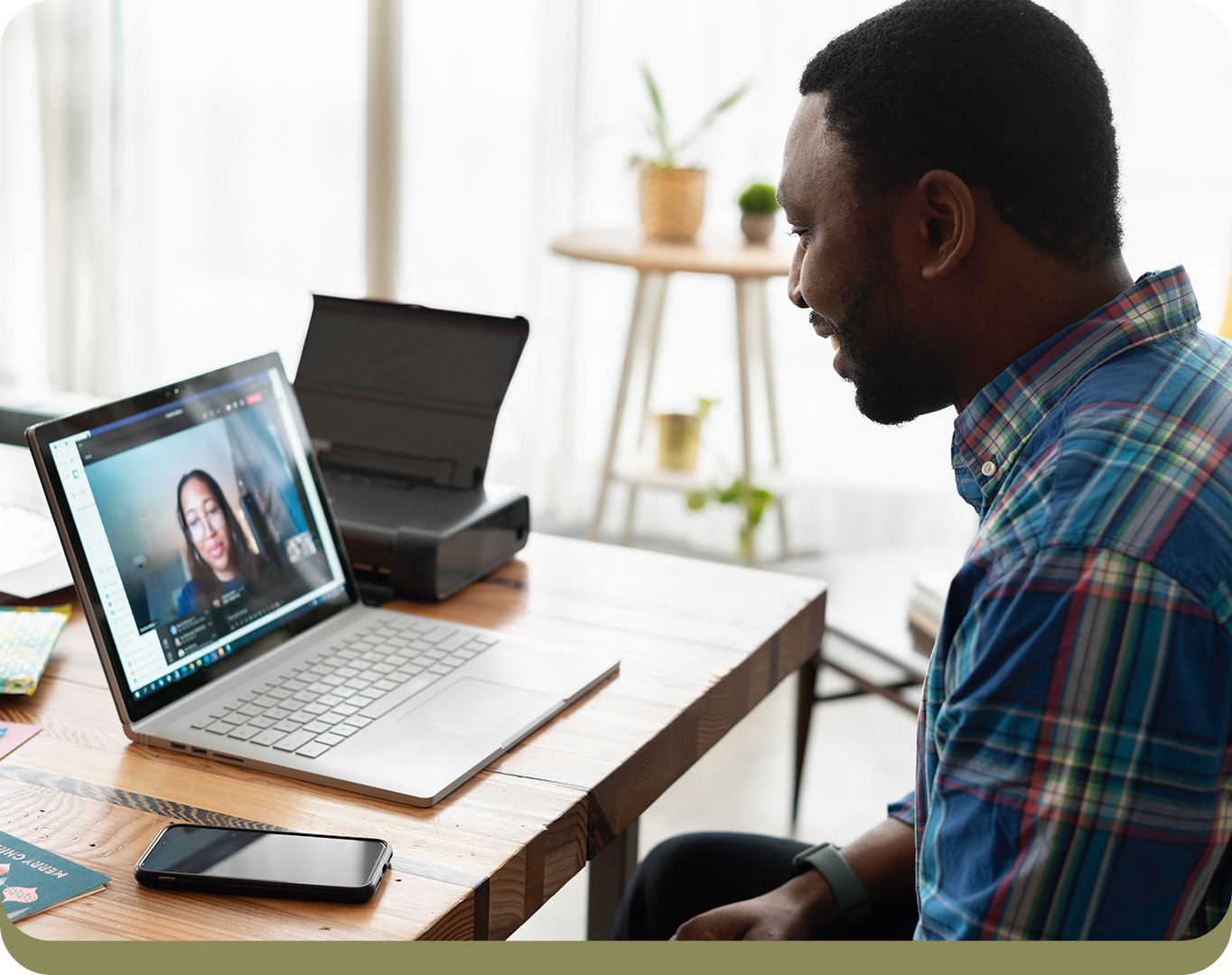 A man in a blue plaid shirt sits at a desk engaged in a video call with a woman displayed on his laptop screen.