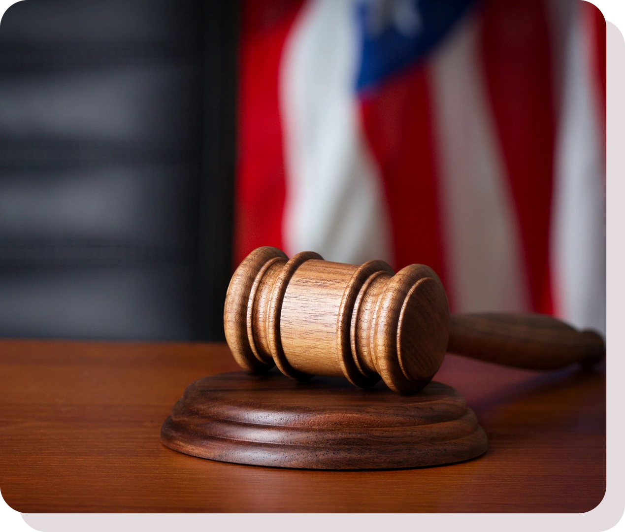 A wooden gavel rests on a sound block on a judge's desk, with a blurred American flag in the background.