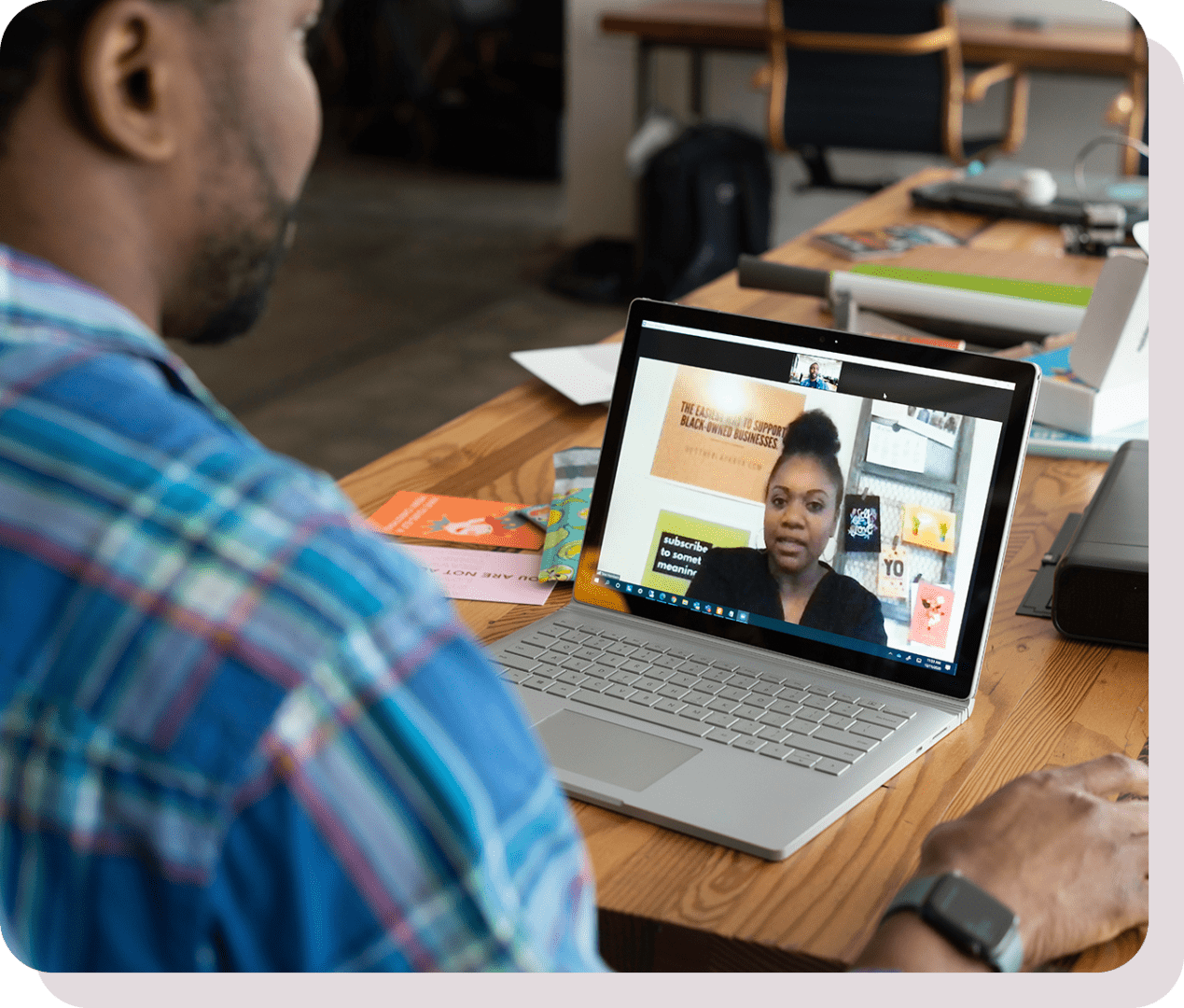 A person wearing a plaid shirt participates in a video call with another individual on a laptop in a workspace.