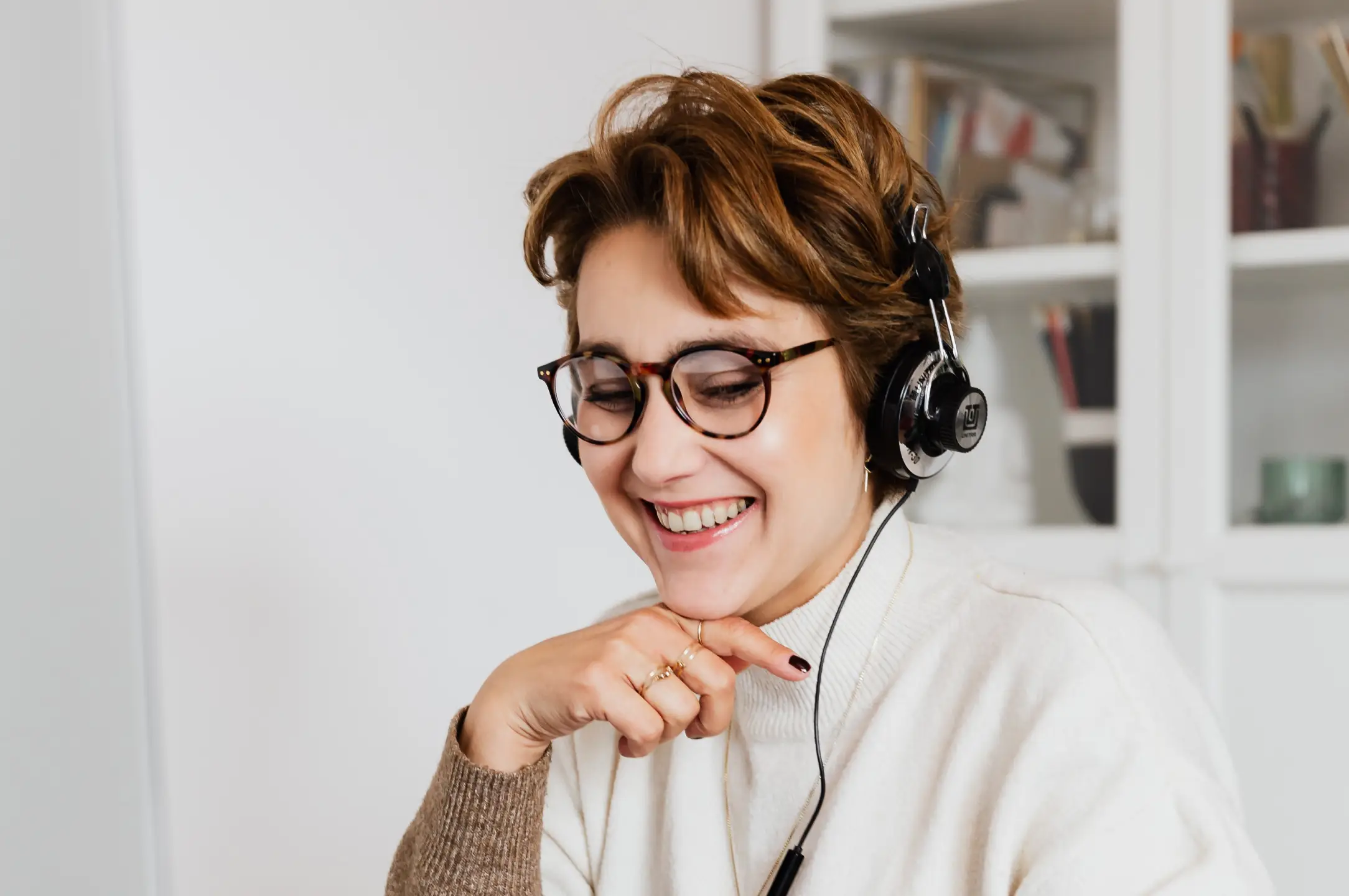 A person with short hair and glasses smiles while wearing headphones, sitting in the PI Online Training Office, a well-lit room with shelves in the background.