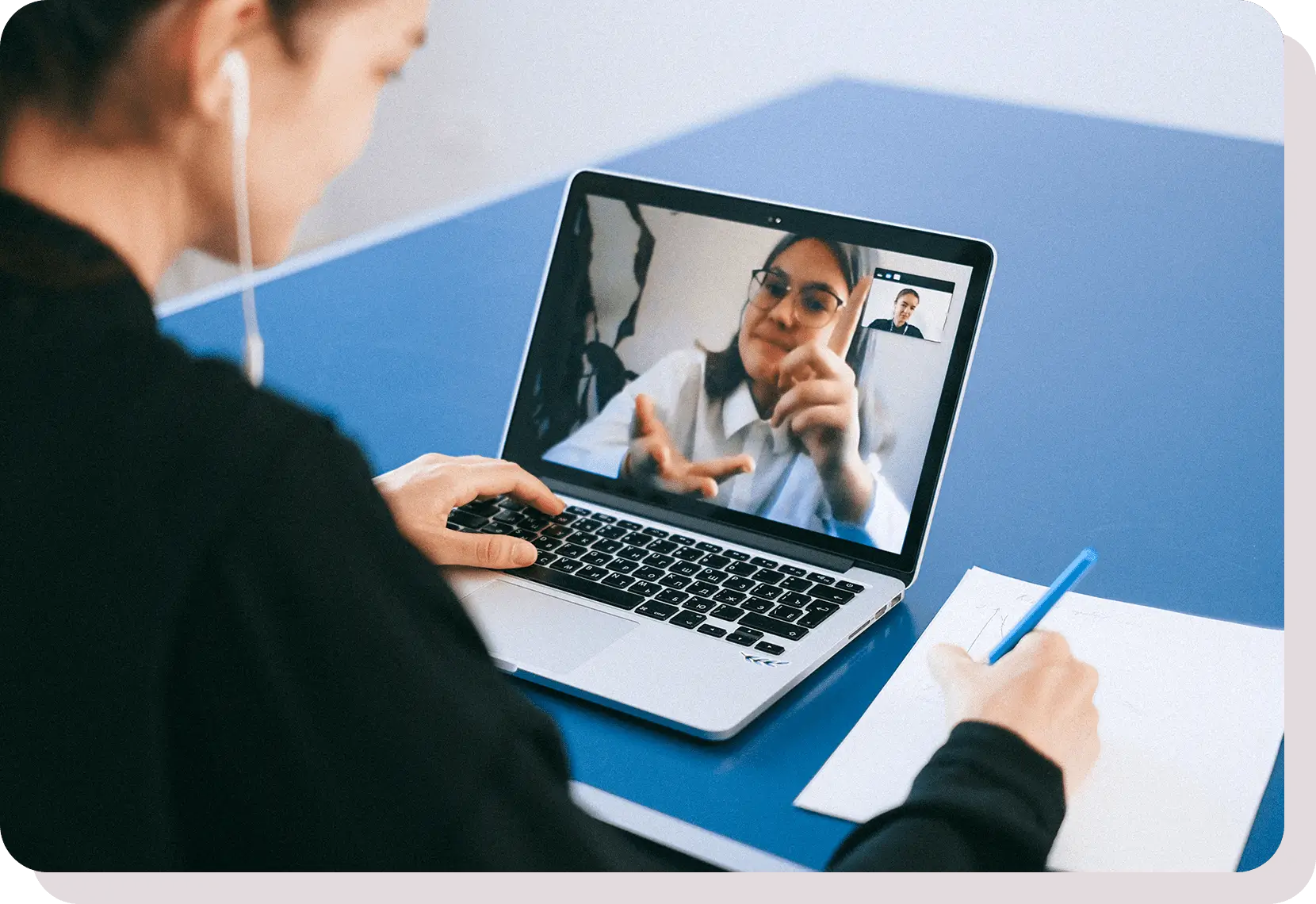 A person wearing earphones is engaged in a video call on a laptop, taking notes with a blue pen on a piece of paper.